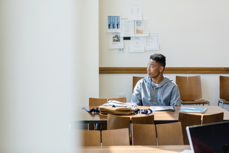 Young man sitting at table in classroom, thoughtful expression. Casual attire, backpack present.